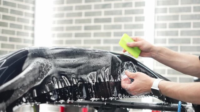 Professional auto detailer specialist applying a clear paint protection film PPF on a glossy black car. Male technician worker smoothing a wet polyurethane protective wrap with a green squeegee rakel.