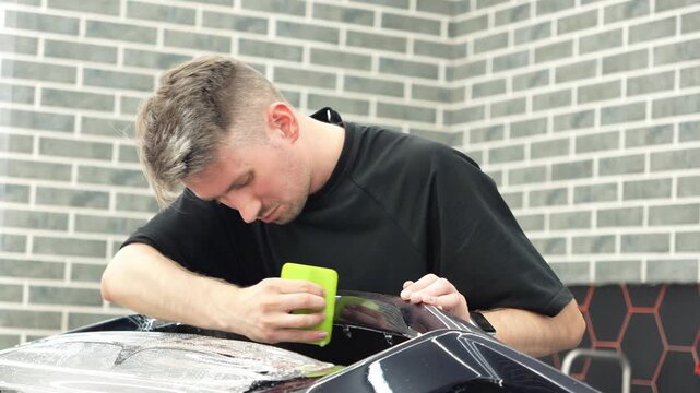 Professional auto detailer specialist applying a clear paint protection film PPF on a glossy black car. Male technician worker smoothing a wet polyurethane protective wrap with a green squeegee rakel.