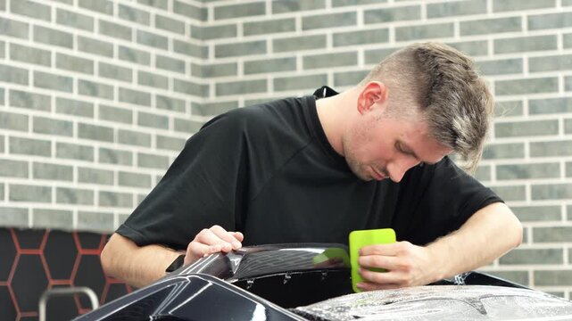 Professional auto detailer specialist applying a clear paint protection film PPF on a glossy black car. Male technician worker smoothing a wet polyurethane protective wrap with a green squeegee rakel.