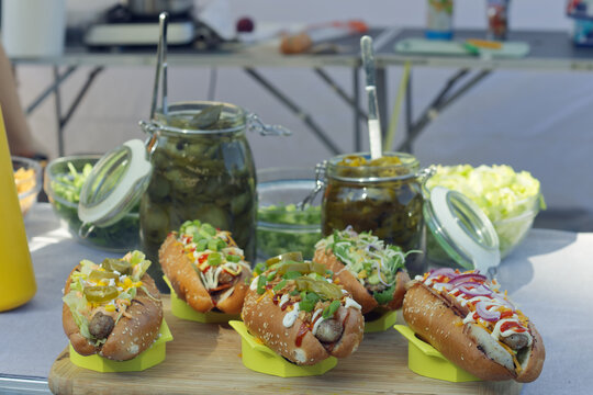 Close-up of gourmet hot dogs with fresh toppings on a rustic wooden board, pickle jars blurred behind at a sunlit Naplavka market stall in Prague.

