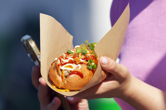 Woman holds a loaded street food bun with mayonnaise, chili sauce and crispy onions at the popular Naplavka farmers market in Prague, phone ready to share the moment.