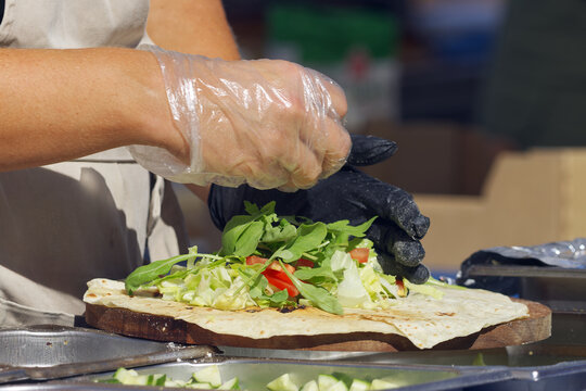 A gloved hand sprinkles crumbled cheese over a fresh flatbread wrap piled with lettuce, arugula, tomato and diced savory toppings at a Naplavka market stall in Prague.