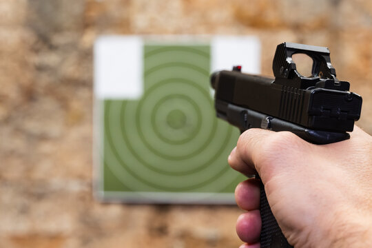 Close-up first-person perspective of a shooter holding a modern pistol with a reflex red dot sight aiming at a paper target in a shooting range.