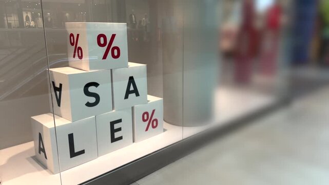 Sale window display uses stacked cubes with bold percent signs and sale letters behind glass. Soft reflections and blurred shoppers in background suggest retail promotion in bright storefront