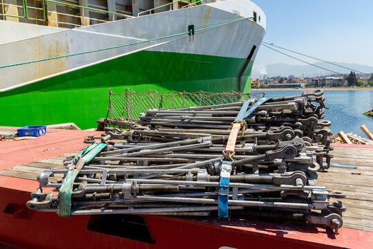 Heavy-duty steel turnbuckles and tensioning hardware secured with cargo straps on a ship deck, with a large cargo vessel moored in the background.