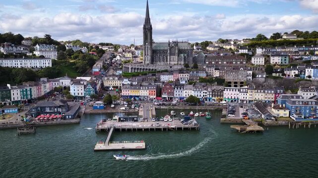 Aerial footage of coastal town of Cobh in Ireland with neo Gothic St Colman Cathedral dominating the hillside above colorful historic houses, waterfront promenade and harbor pier.
