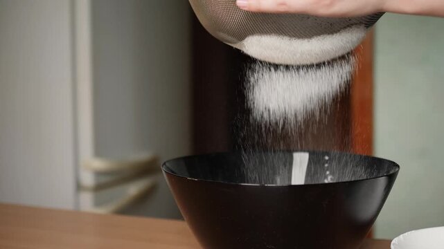Close up view of hands sifting white wheat flour through metal sieve into black bowl for baking dough