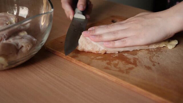 Close up view of hands slicing raw chicken breast fillet horizontally with sharp knife on wooden cutting board