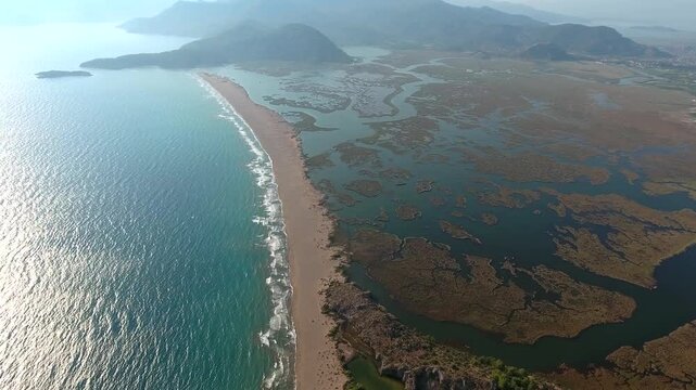 Aerial view shows Iztuzu Beach and Dalyan Delta lagoons in Mugla Turkey beneath blue sky. Sandbar channels weave through marshland, creating coastal wetland labyrinth beside the sea.