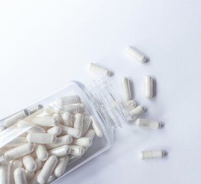 White medicine capsules and tablets spilling from an open transparent bottle on a white background. Medical, pharmacy, healthcare, vitamins and dietary supplements concept with copy space.
