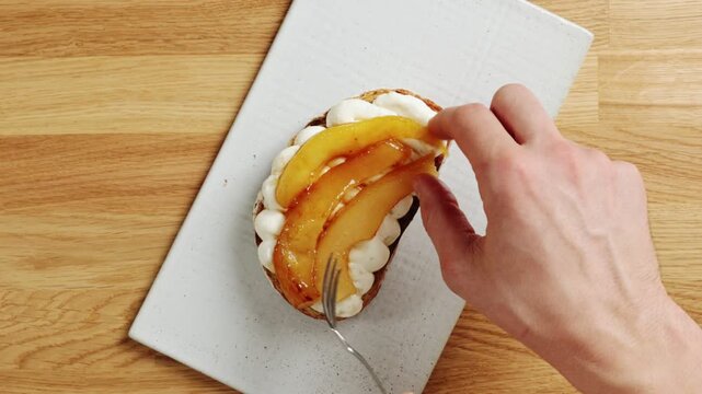 Hands arrange caramelized pear pieces on ricotta toast with fork. Food stylist creates clean composition for food photography. Finished brunch plate suits social media, restaurant menu or recipe blog.