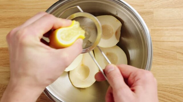 Hand squeezes lemon juice through sieve over pear halves in bowl. Citrus juice keeps fresh fruit pale and prevents browning. This kitchen trick prepares pears for salad, tart, dessert or lunchbox.