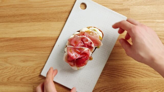 Hands show prosciutto ricotta toast to camera in timelapse. Food blogger presents creamy cheese and cured ham on crisp bread. This brunch recipe suits social media, cooking tutorial or cafe review.