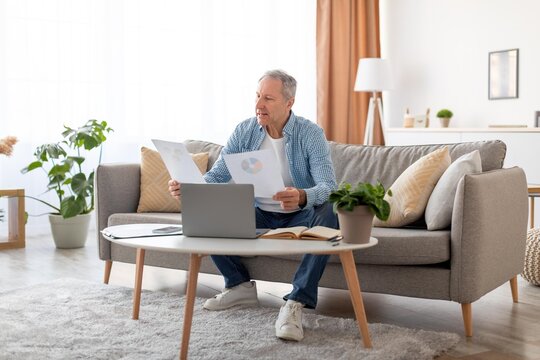 Focused Mature Caucasian Man Reading Business Reports While Sitting On Couch In Home Office, Engaging With Financial Data On Paper, Laptop On Table, Indoor Environment. Business Concept.
