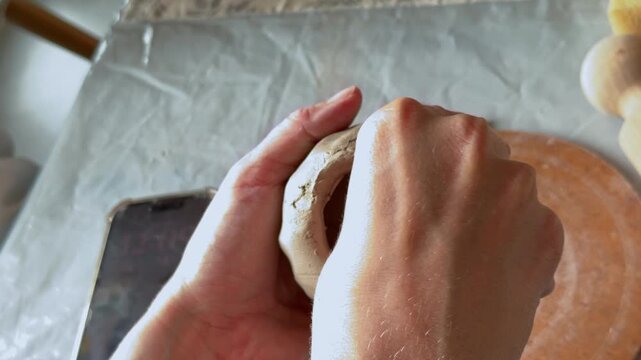 A person molds a small clay vessel with both hands on a covered worktable, hollowing the center to form a rounded pot in a handmade pottery session.
