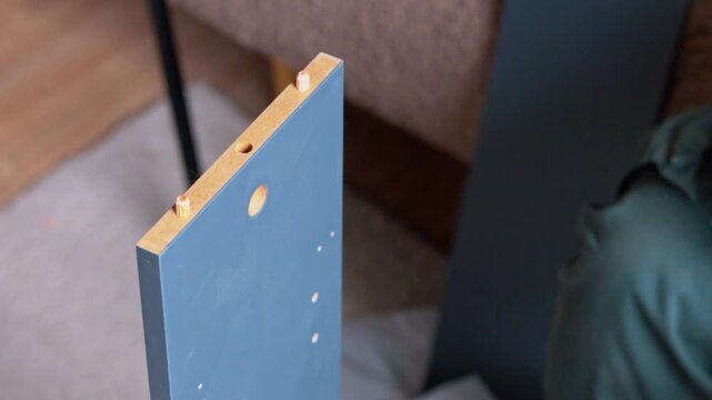 Person assembling furniture piece by inserting wooden dowels into pre-drilled holes of a blue panel, with additional panels and a soft surface visible in the background