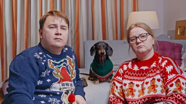 An adult man and woman in Christmas sweaters sit on a sofa with a black and tan dachshund dog wearing a knitted sweater between them, all facing the camera for a festive family portrait.