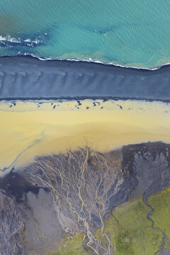 Aerial view of a glacial river delta with intricate braided river channels, black sand deposits, and turquoise coastal waters, Iceland.