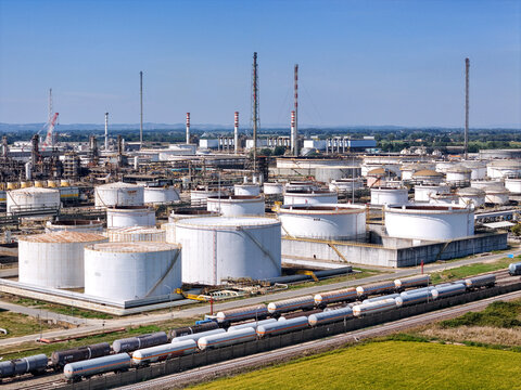 Aerial view of the Eni oil refinery featuring large white storage tanks, industrial infrastructure, and a freight train loaded with tanker cars, Sannazzaro de' Burgondi, Lombardy, Italy.