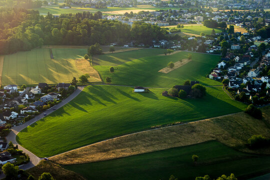 Aerial view of lush green fields and meadows surrounded by suburban houses, trees, and winding roads under the warm light of the golden hour in Zurich, Zurich, Switzerland.