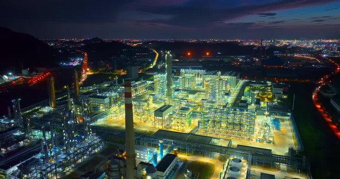 Aerial view of an illuminated chemical refinery plant at night, showing tall distillation columns, complex pipeline networks, and bright industrial lighting.