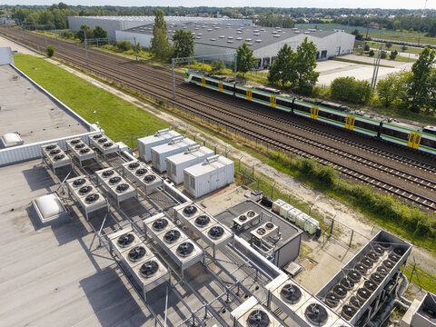 Aerial view of industrial cooling units and HVAC systems on a data center rooftop with a passenger train passing on nearby tracks in Poland.