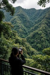 Obraz premium Young woman photographer uses digital camera to take photo of green mountains. Female traveler explores forest landscape, scenic wilderness. Hobbyist captures nature views from wooden balcony in