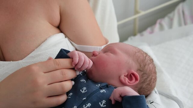 Adorable newborn baby boy in a blue onesie sleeping peacefully in his loving mother's arms while she gently holds his tiny hand, representing a beautiful moment of family bonding and care