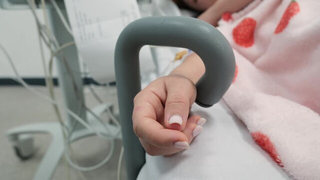 Close-up of a woman's hand trembling involuntarily on a hospital bed railing during medical treatment, with an IV line and medical equipment visible in the background