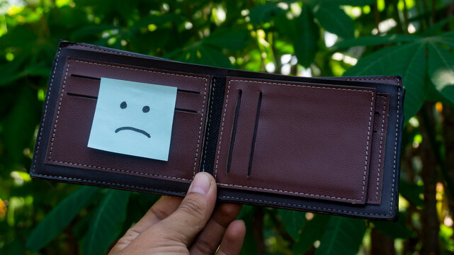 Brown wallet displayed outdoors with a blue sad face sticky note in the card slot, representing financial stress, empty wallet, debt, or personal budgeting concept.