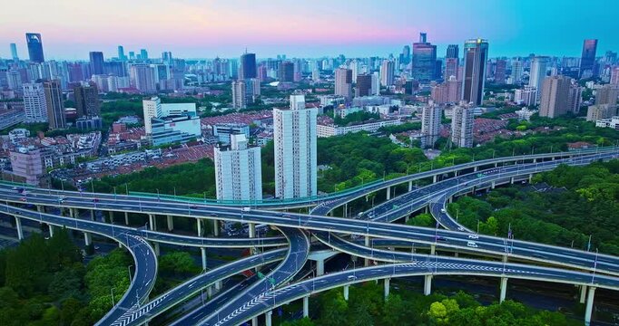 Elevated view of a multi-level highway interchange surrounded by lush green trees, with Shanghai city skyscrapers under soft sunset sky.