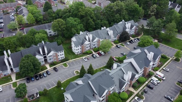 Aerial drone footage tracking over a suburban apartment complex. Features multi-story residential buildings with gray roofs, parking lots filled with cars, and lush green trees in soft daylight.