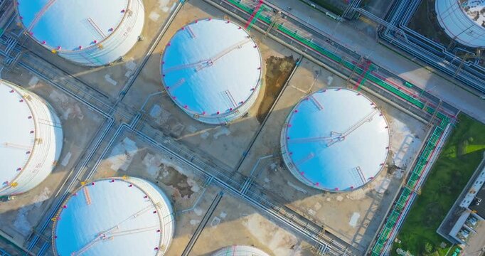 Aerial view of large circular oil storage tanks and complex pipeline systems in an industrial refinery complex illuminated by bright sunlight.