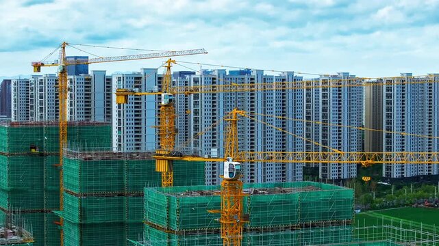 Yellow tower cranes operate over apartment blocks wrapped in green safety netting under a bright cloudy sky in natural daylight.