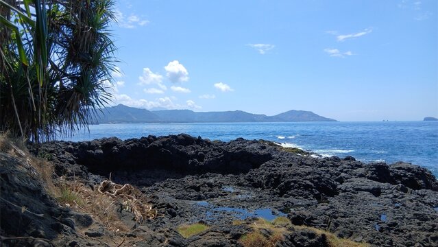 DENPASAR, BALI,INDONESIA september,05,2026:Black volcanic rock coastline at Padang Bai Beach, Bali with mountain view and clear blue sea