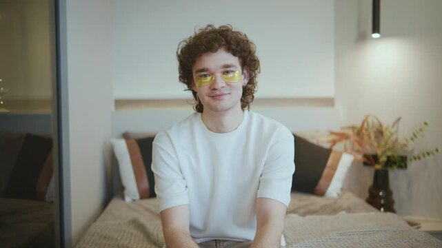 A young man sits on a sofa in a cozy apartment, smiling warmly at the camera while wearing a simple T-shirt, with skincare patches under his eyes