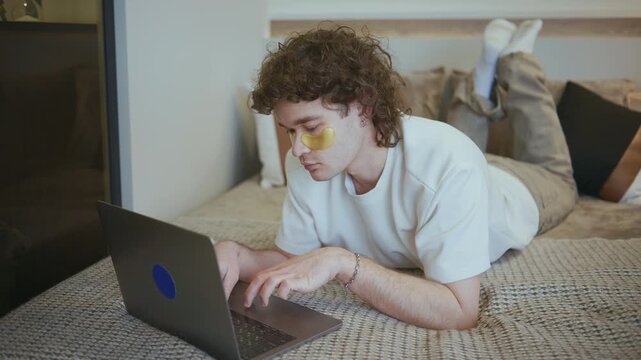 A young man is lying on his stomach on a bed, using a laptop. He is wearing yellow under-eye patches. The room has simple decor, creating a relaxed atmosphere.