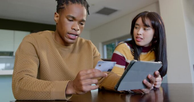 Diverse couple at dining table, man showing payment card to tablet folio case to pay, hugging