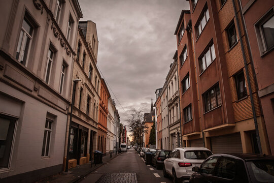 A narrow residential street in the Nippes district of Cologne, germany, framed by multistorey apartment buildings with cars parked along both curbs under an overcast evening sky.