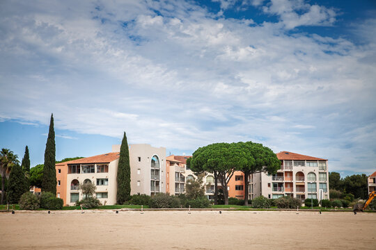 Holiday residence on the French Riviera, showing Mediterranean-style apartment buildings with terracotta roofs, balconies and landscaped greenery beside a sandy open area in southern France.