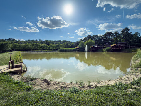 Scenic wide-angle view of a calm lake with a water fountain, wooden cabins along a forested shore and a sluice gate on the grassy bank beneath a bright blue sky with white clouds.