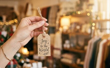 Obraz premium Woman holding a Christmas gift tag with Joy to the World text in a boutique shop with festive holiday decor and warm bokeh lights for seasonal greeting concepts