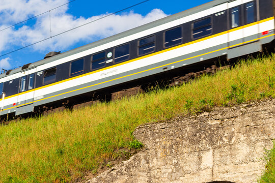 Modern passenger commuter train speeding along elevated track against blue sky, motion blur on metal railcar passing grassy hillside on a sunny summer day