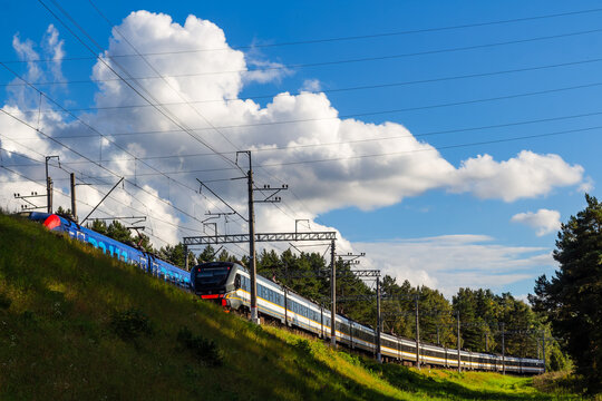 Two modern commuter trains moving in opposite directions along parallel railway tracks through a scenic green landscape under a bright blue sky with fluffy white clouds