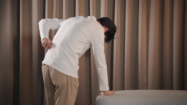 Asian man rubbing lower back while standing by sofa experiencing sharp spinal pain and muscle stiffness indoors.