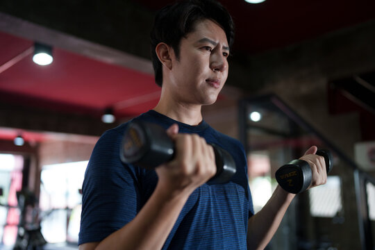 Young Asian man performing dumbbell bicep curl exercise during strength training workout at modern fitness gym