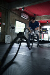 Obraz premium Young Asian man performing intense battle rope exercise in modern fitness center gym interior