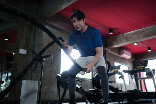 Young Asian man performing battle rope exercise at modern fitness gym during intense cardio training session