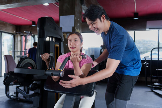 Young Asian personal trainer guiding adult woman through arm exercise on weight machine at modern fitness center