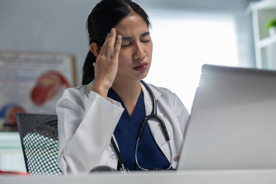 Asian woman doctor experiencing headache while using laptop in medical office, expressing fatigue and workplace stress during demanding healthcare shift